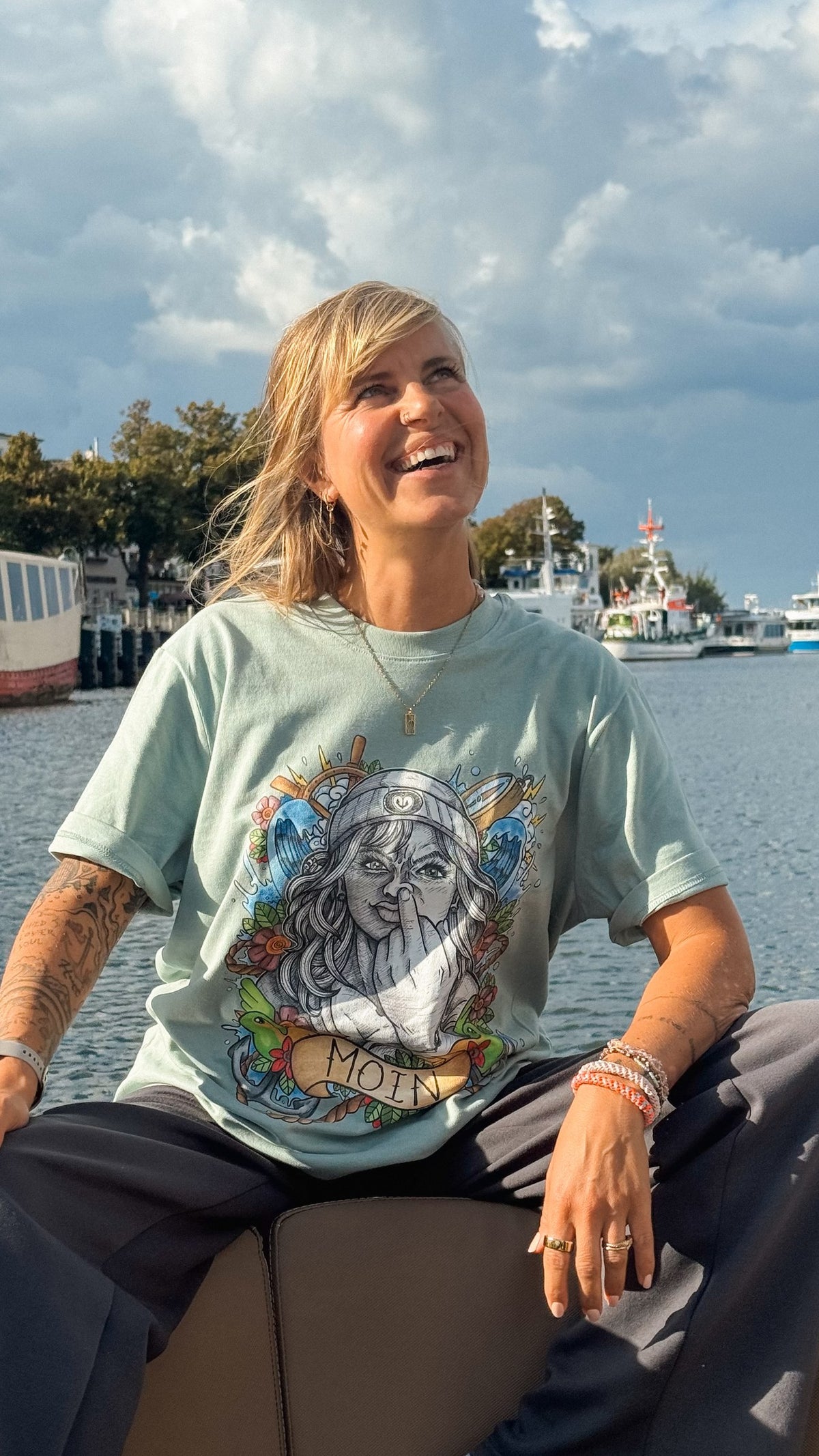 Sitzende Frau auf einem Boot im mintfarbenen T-Shirt mit farbigem Moin-Motiv vor dem Hafen.