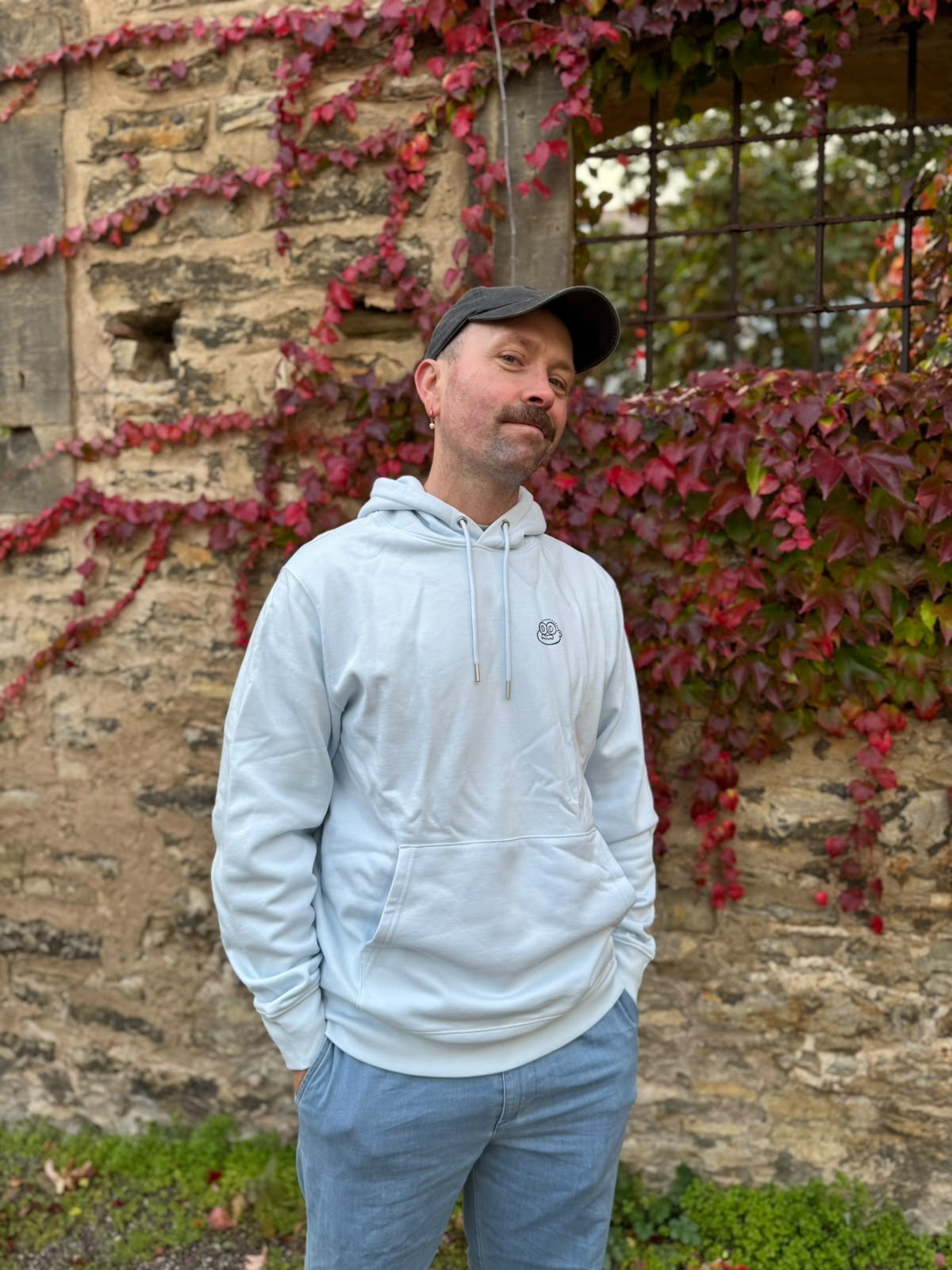 Mann mit Cap trägt hellblauen Hoodie mit kleinem rundem Frontprint vor einer Steinmauer mit rotem Herbstlaub