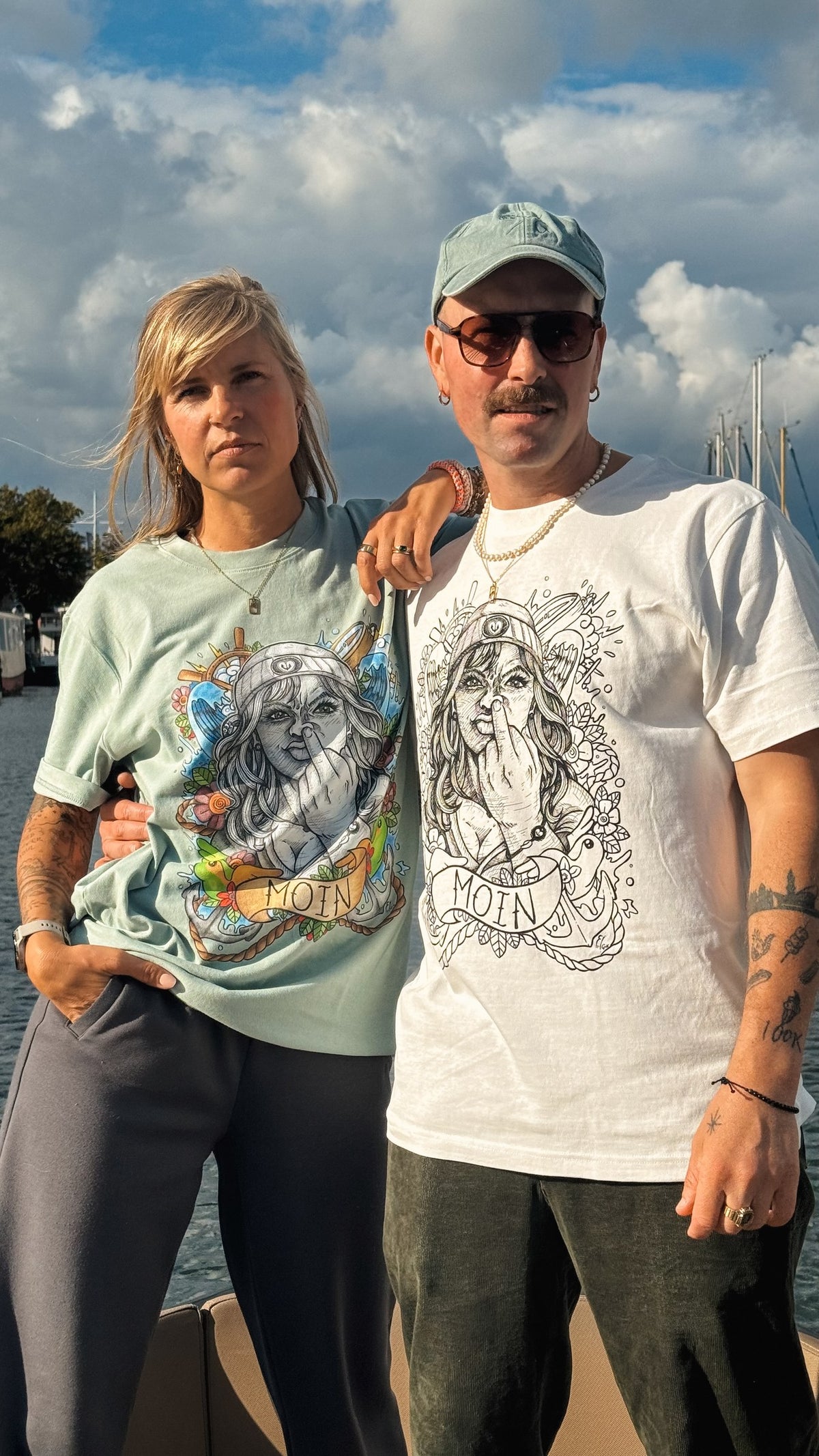 Zwei Personen am Wasser mit mintfarbenem und weißem T-Shirt, beide mit Moin-Motiv auf der Vorderseite.