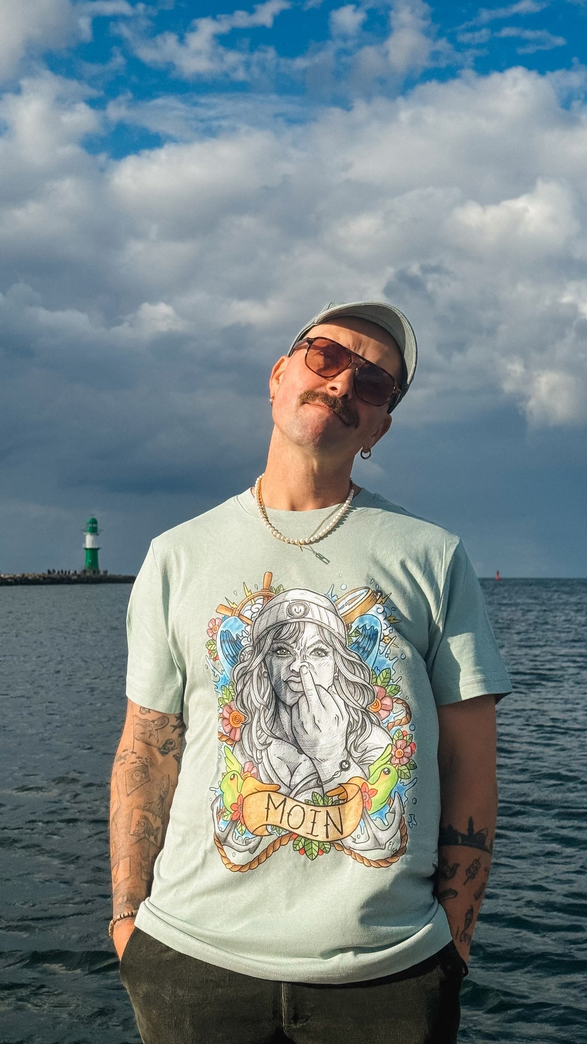 Mann vor der Ostsee im mintfarbenen T-Shirt mit großem farbigen Moin-Motiv.