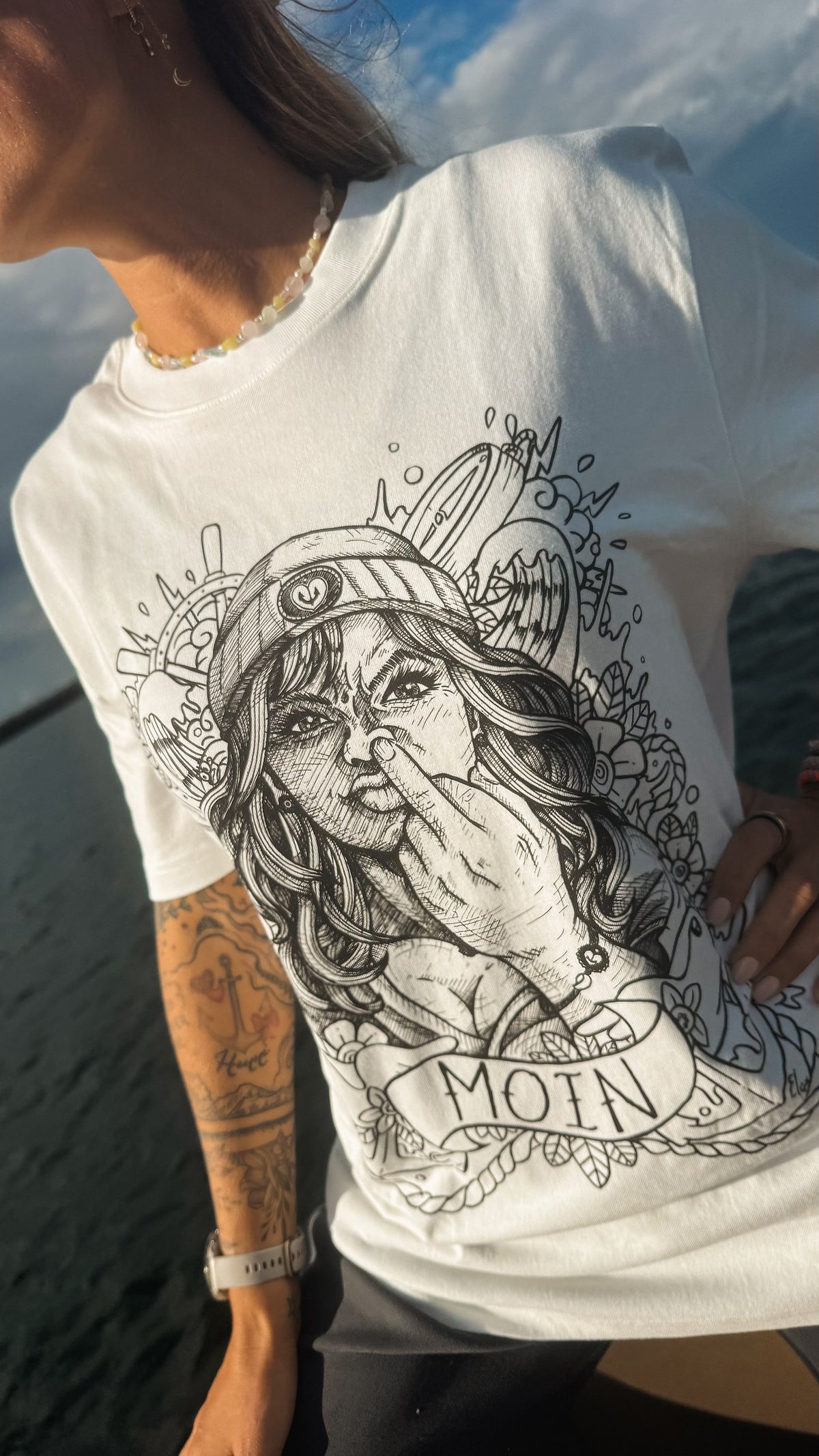 Detailansicht des weißen Shirts mit gezeichneter Kapitänin, Seemannsmütze und MOIN-Banner.